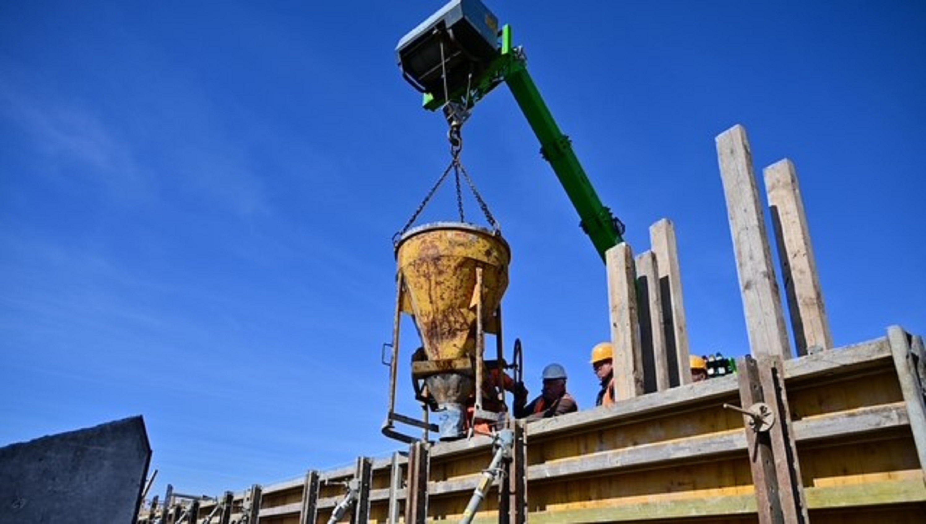 Bauarbeiter füllen mit einem Kran und gelbem Trichter Beton in eine Schalung, blauer Himmel im Hintergrund.








                                  
